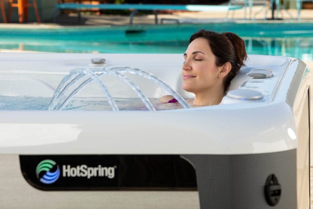 Woman relaxing with eyes closed in a white HotSpring hot tub by a pool, jets spraying water nearby.