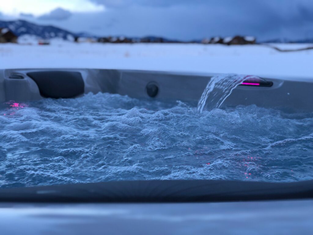 Close-up of a hot tub with bubbly water and a pink-lit edge against a snowy landscape and blue sky behind.