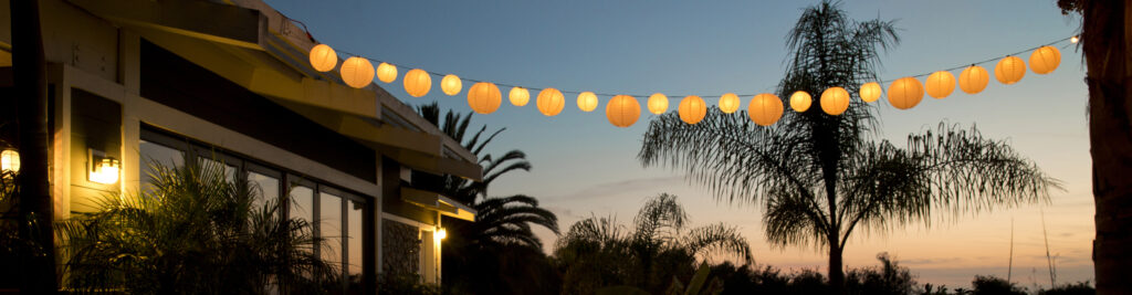 Evening exterior of a modern house with orange lanterns strung overhead and palm trees in the yard.