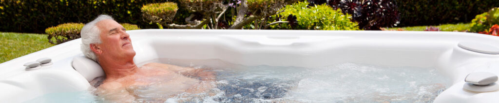 Older man relaxing in a white outdoor hot tub with bubbling water, eyes closed, surrounded by greenery.