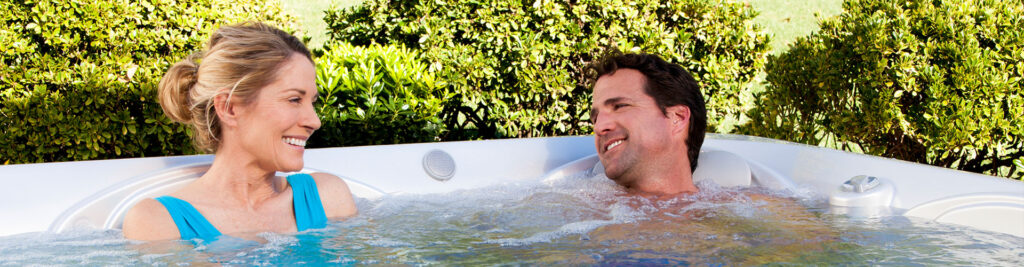 Couple relaxing in a white hot tub outdoors, smiling at each other with green bushes in the background.