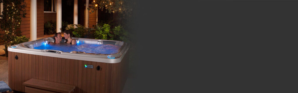 Couple relaxing in an outdoor wooden hot tub at night, with string lights and greenery around.