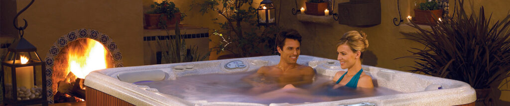 A couple sits in an outdoor hot tub at night, smiling as they relax by a lit fireplace and lanterns nearby.
