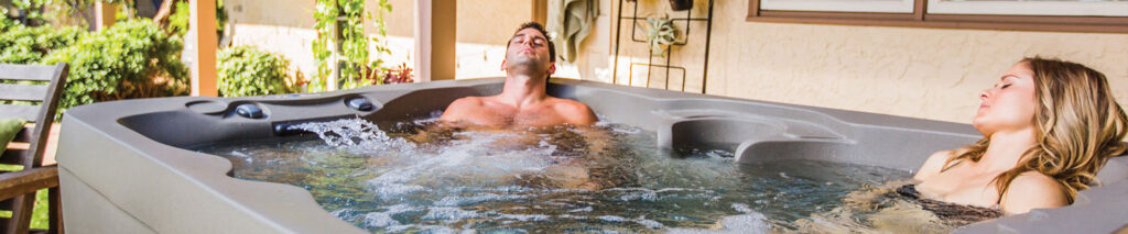 Couple relaxing in a gray outdoor hot tub in a sunny backyard, leaning back and enjoying the jets.