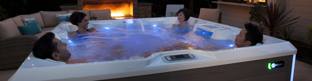 Four adults relax in a glowing, blue-lit outdoor hot tub at night beside a lit fireplace and patio seating.