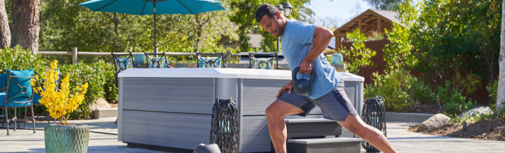 Man in a blue shirt and gray shorts lunges with a kettlebell outdoors on a sunny patio.