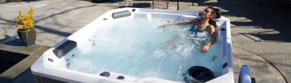 Woman relaxing in a white outdoor hot tub in a sunny patio, blue swimsuit and bubbles visible in the water.