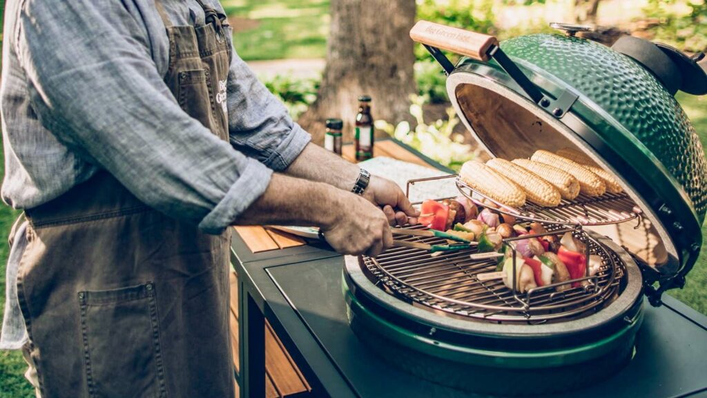 Person in a denim apron turns vegetable skewers on a round green outdoor grill.