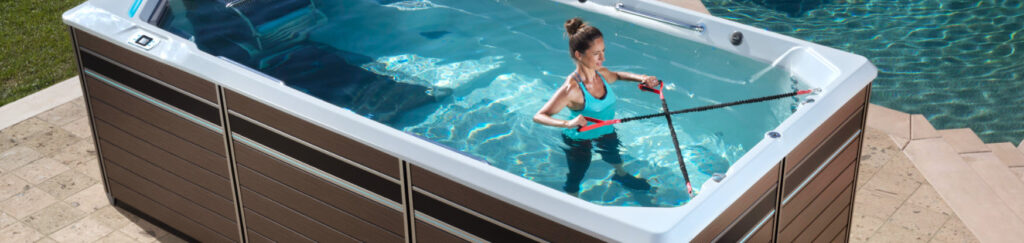 Woman in a turquoise swimsuit uses red-and-black resistance bands to train in a rectangular outdoor hot tub by a pool edge