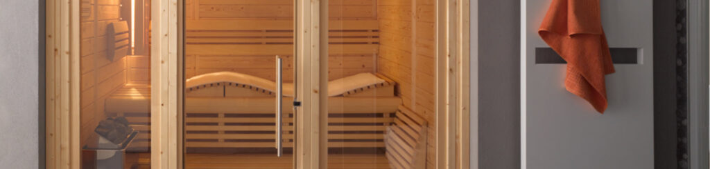 Glass-door sauna interior with wooden benches and a heater with stones; an orange towel hangs nearby on a panel outside the door.