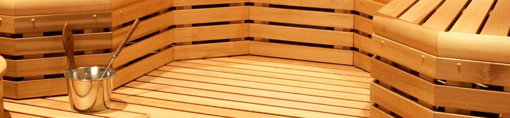Inside a wooden sauna with tiered benches and a metal bucket with a ladle on the floor/seat edge.