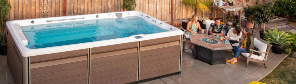 Outdoor backyard scene with a brown hot tub on the left and five people around a square fire pit table on a concrete patio.
