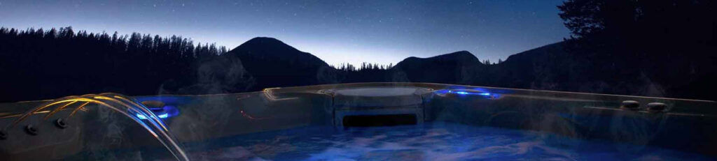 Night photo of an illuminated hot tub overlooking a dark mountain landscape under a starry sky with mist rising from the water.