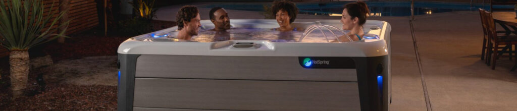 Four friends soak in a modern HotSpring hot tub at night by a pool, smiling and chatting.