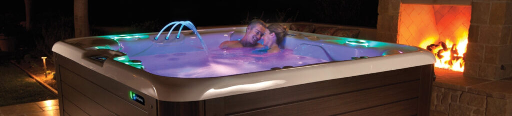 Couple relaxing in an illuminated outdoor hot tub at night beside a lit stone fireplace.