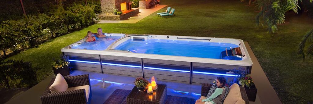 Nighttime backyard scene with a illuminated hot tub and two people relaxing inside while a seating area with candles sits nearby.