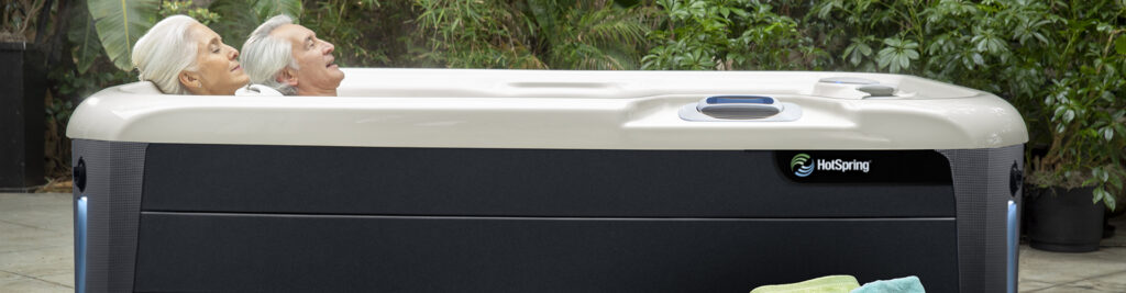 Two elderly people relaxing in an outdoor HotSpring hot tub with a white rim and dark base, surrounded by tropical plants.