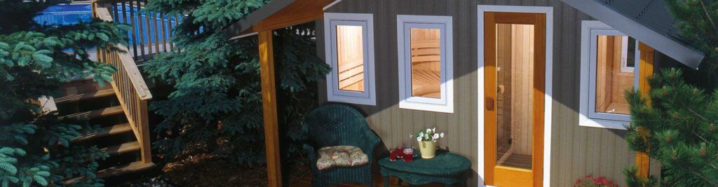 Cozy wooden cabin exterior with a small porch, wicker chair and table of drinks, surrounded by evergreen trees and shrubs.