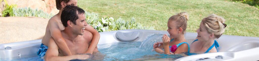 Father and two girls laughing and splashing in an outdoor hot tub on a sunny day.