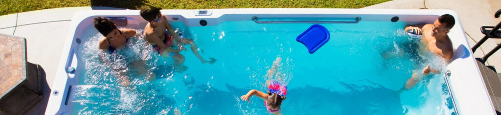 Top-down view of a backyard pool with four people—two adults and two children—playing and splashing, a blue kickboard floating on the water.
