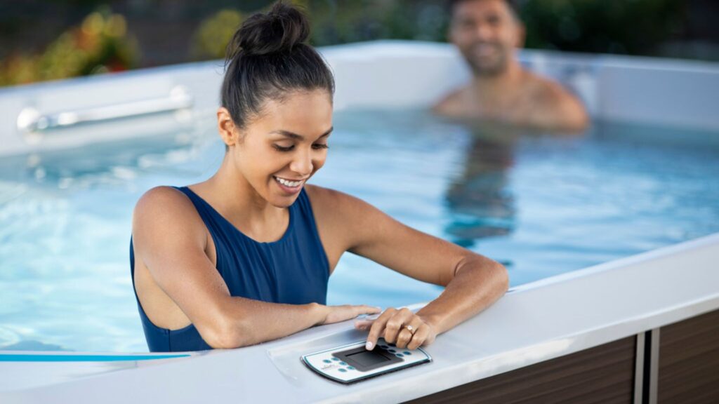 Woman in a blue swimsuit adjusts the hot tub controls at the edge while a man relaxes in the background outside.
