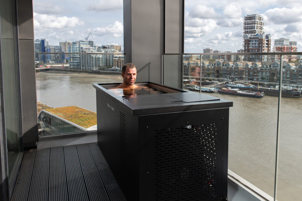 Man relaxing in a rooftop hot tub on a balcony overlooking a river and city skyline.