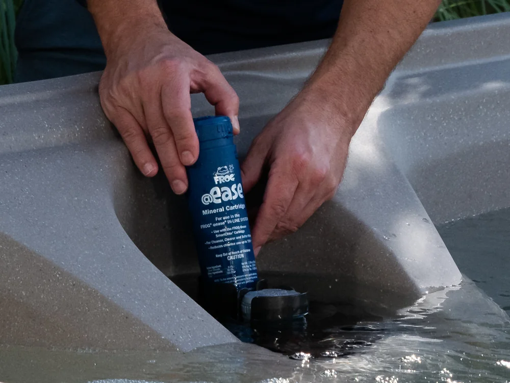 Person inserts a blue Frog @ease mineral cartridge into a spa or pool filter housing while hands rest in water-filled tub liner