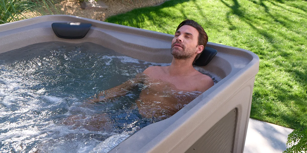Man relaxing in a gray outdoor hot tub with his eyes closed, sunlight on the water and a grassy backyard in the background.