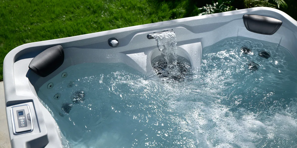 Outdoor hot tub with active jets and a small water feature, built-in control panel on the left and green lawn in the background.