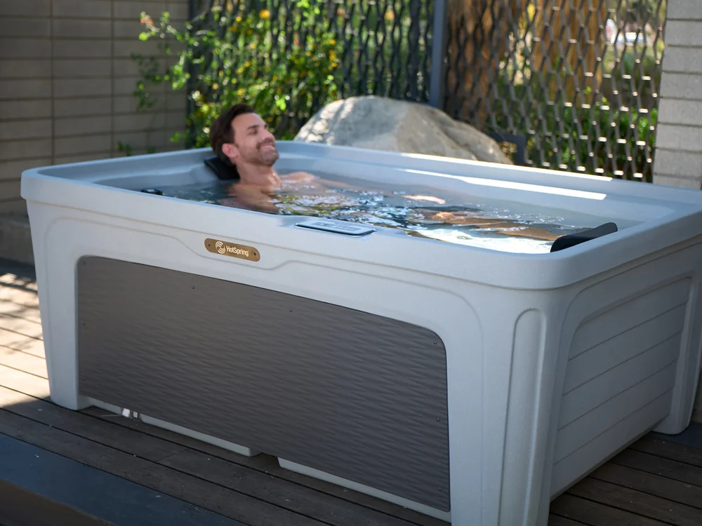 Man relaxing in an outdoor hot tub on a wooden deck, leaning back with eyes closed, sunny garden in the background